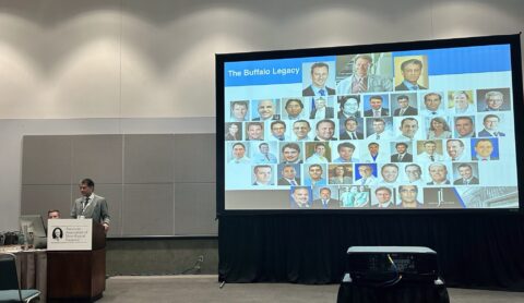 picture of Dr. Siddiqui standing at podium next to big projector screen. PowerPoint slide shown says The Buffalo Legacy with images of UBNS physicians