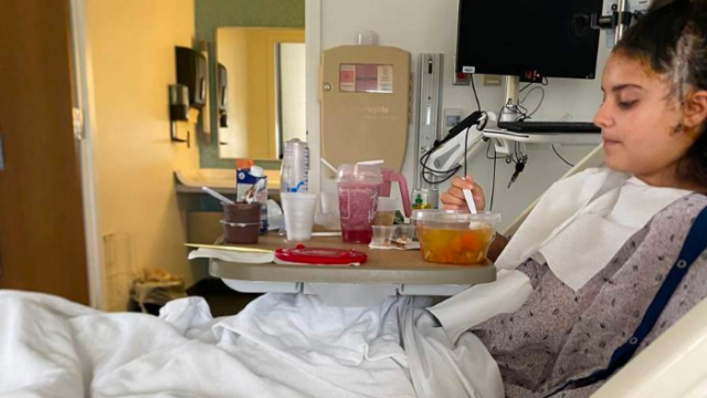 Noa Haroush sits in her hospital bed eating soup and is surrounded by various beverages and pudding cups.